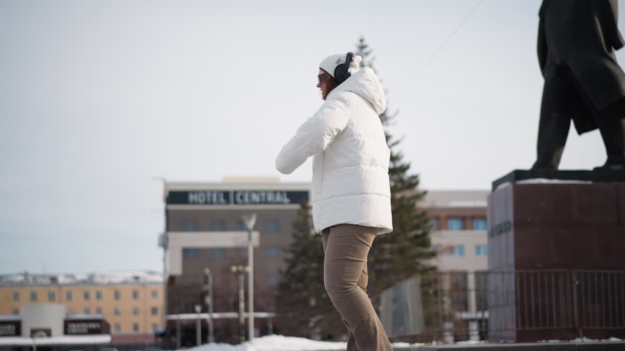 Young lady wearing white winter coat, sunglasses and headset dances joyfully behind statue on paved plaza in snowy park setting with grand building facade and trees under bright cold daylight