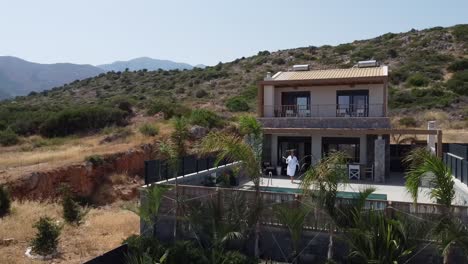 una mujer delgada de cabello oscuro camina a una piscina privada y se quita la bata y se sienta en un traje de baño a la piscina - villa en grecia creta y las palmeras en primer plano se pueden ver