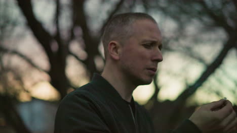 a man in a green jacket stands outdoors at dusk, holding a cigarette to his mouth, deep in thought, with blur view of cars moving from behind