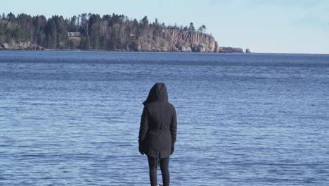 woman in rain jacket stands on the edge of lake superior and looks at the rocky shore and cabins across the water