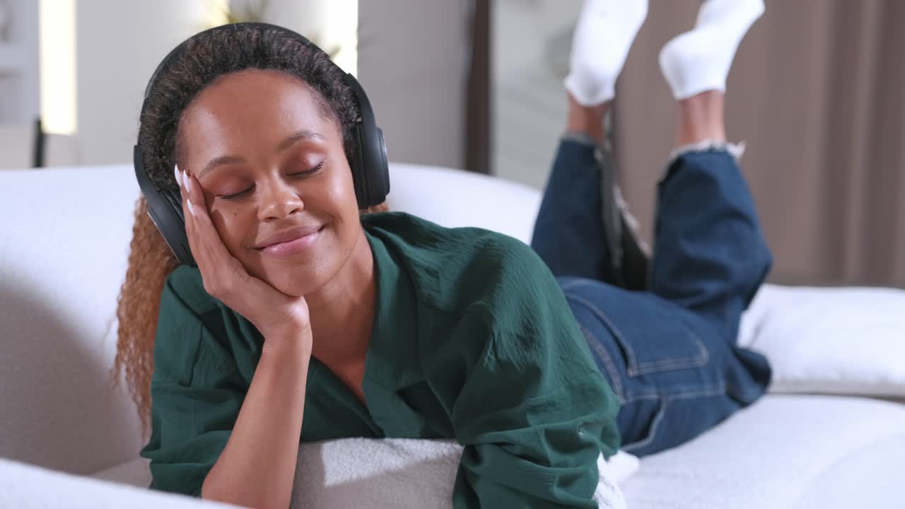 Ung, avslappet afroamerikansk kvinne lytter til musikk på hodetelefoner mens hun ligger på sofaen