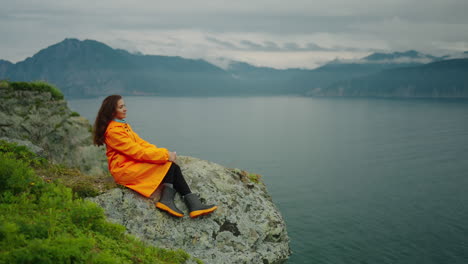 woman in orange raincoat on cliff overlooking lake