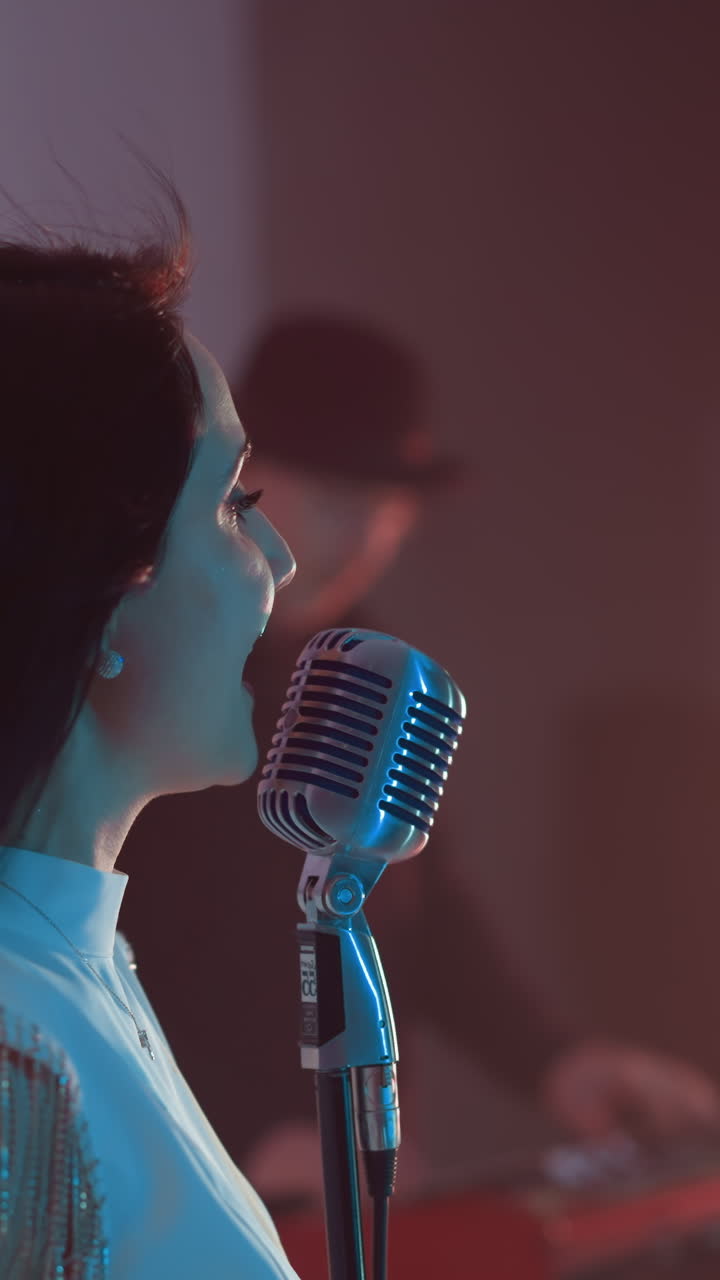 Side view of a singer in a white gown singing and lifting her hands up, with a blurred keyboardist in the background. The scene is set against a soft red and blue background