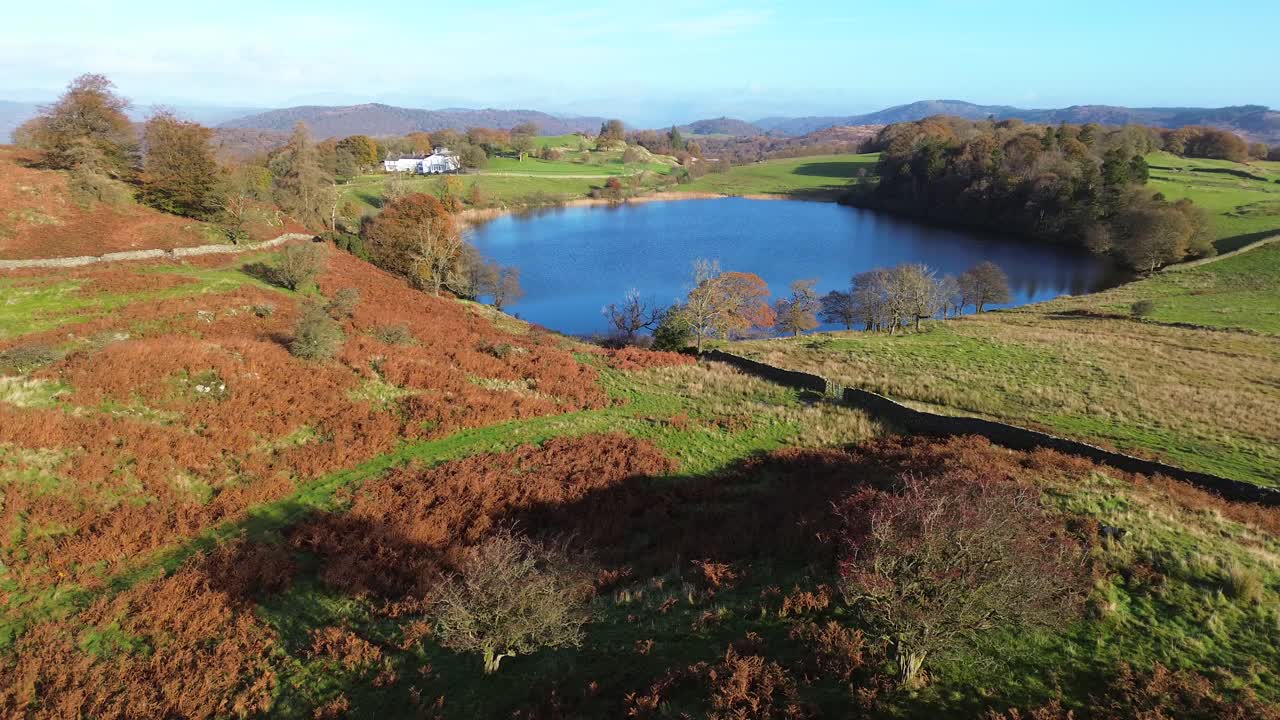 Drone cinematografico che sorvola colline autunnali verso una tranquilla residenza del Lake District e un lago blu