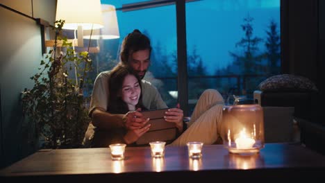 romantic evening: a guy and a girl are sitting on a devan in a room lit by candles and looking at the tablet screen. room overlooking the evening coniferous forest