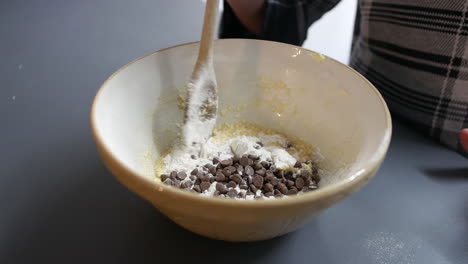 a girl mixing chocolate chips into cookie dough cake mixture in a bowl with a wooden spoon