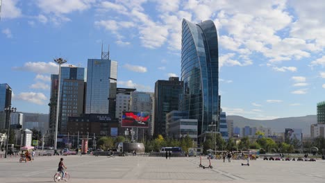 under the bright sun, people gather at sukhbaatar square, with the damdin sukhbaatar statue and modern structures, including the blue sky building, visible in the background in ulaanbaatar, mongolia.
