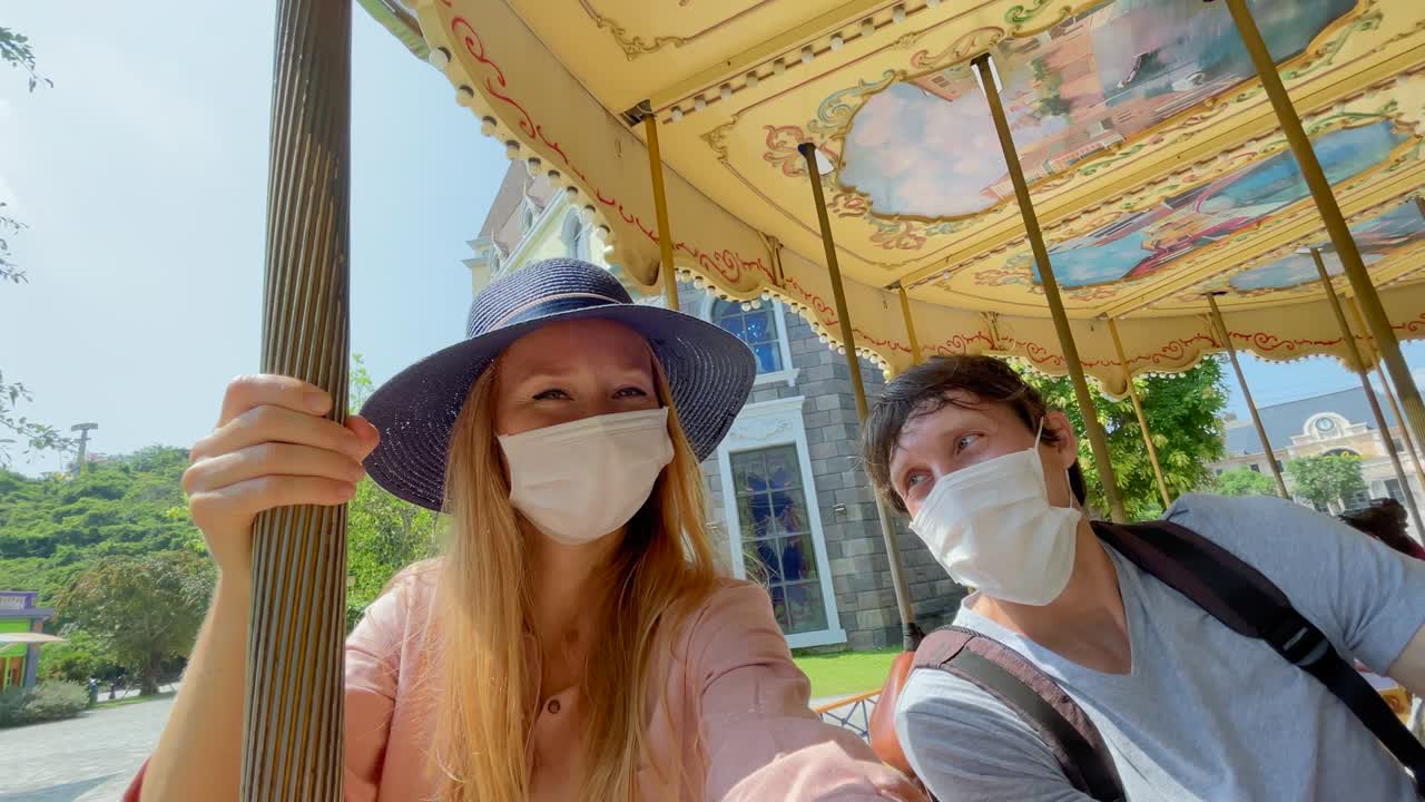 슬로우모션 촬영: 보호 마스크를 착용한 젊은 커플이 카루셀을 타고 팬데 사이에서 공원을 방문합니다.