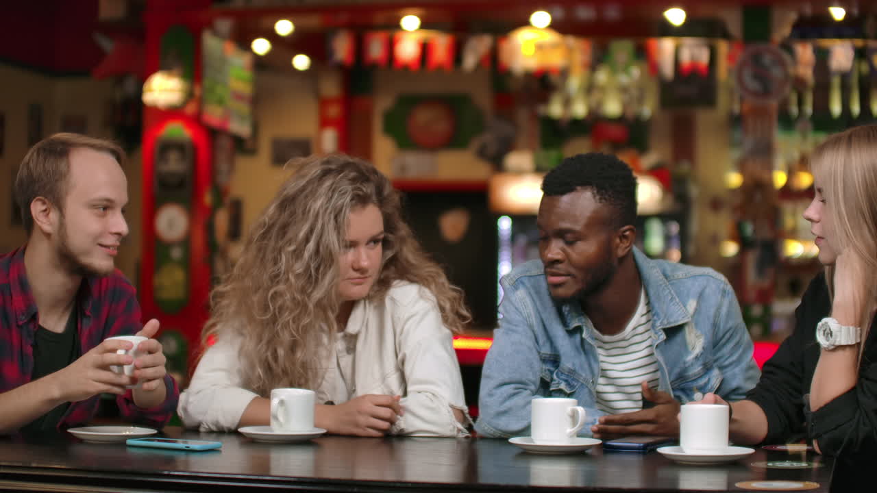 다양한 행복한 친구들이 함께 기기와 함께 재미있는 온라인 동영상을 스마트폰에서보고 있습니다. 다문화 젊은 친구들이 웃고 전화를 보고 이야기하고 모임에서 모바일 앱을 사용합니다. 테이블에 앉으십시오.