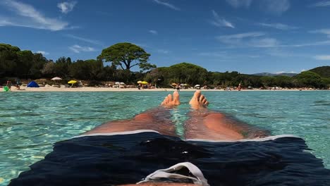 Personal-perspective-and-low-angle-of-man-legs-and-feet-relaxing-while-floating-on-seawater-at-Santa-Giulia-beach-in-Corsica-island,-France