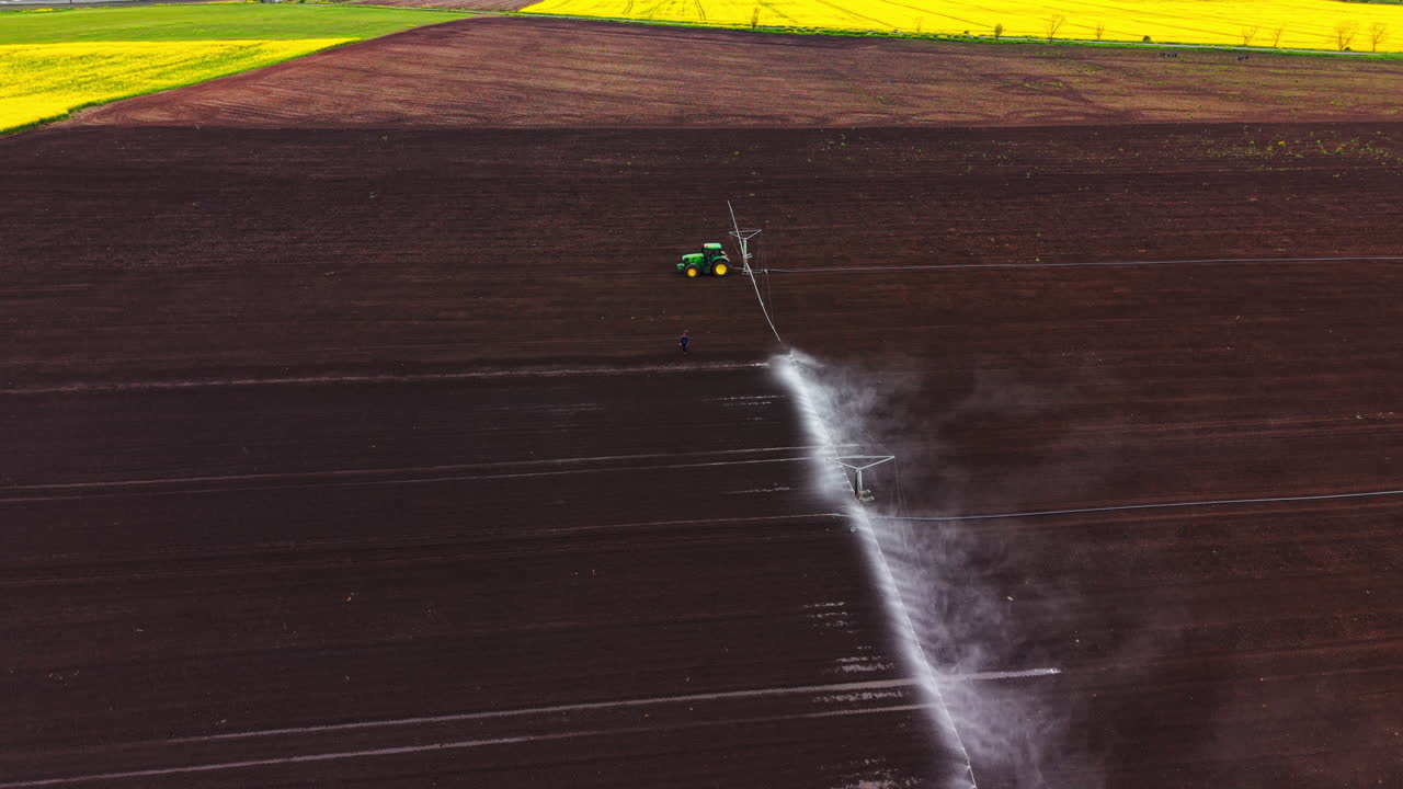 Vista aerea di un trattore verde che traina un'ampia irroratrice per irrigazione attraverso un campo marrone scuro, con un lavoratore in piedi nelle vicinanze e un bordo del campo giallo.