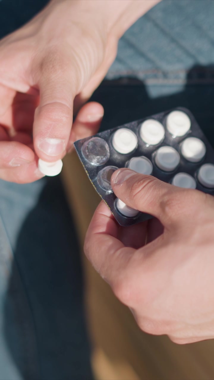 Close up of hands opening blister pack and removing white pills focusing on action. Hands manipulate black plastic packaging containing round tablets under bright light with soft shadows