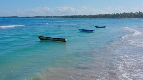 Aerial-drone-shot-of-three-fishing-boats,-waves-on-the-turquoise-blue-beach,-radiant-sun