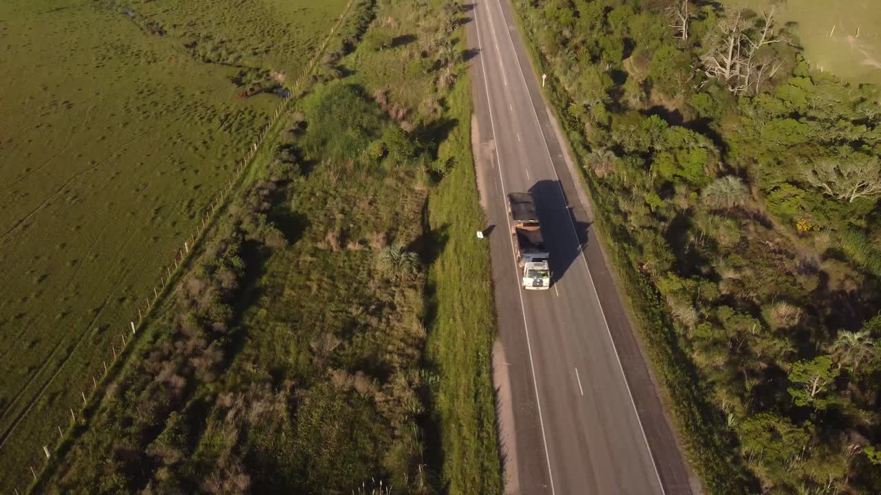 Toma aérea de seguimiento de un camión industrial circulando por una carretera rural asfaltada en Uruguay durante el atardecer