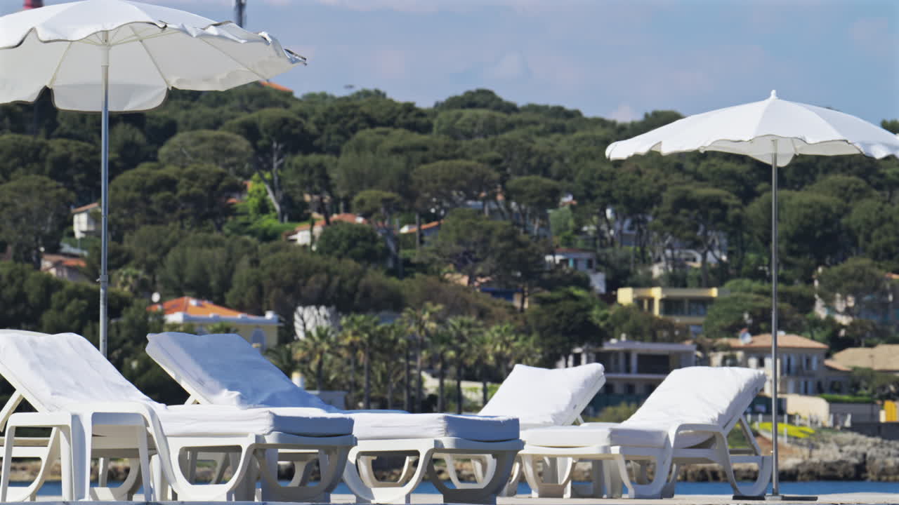 White beach loungers and sun umbrellas with green trees and villas on the background