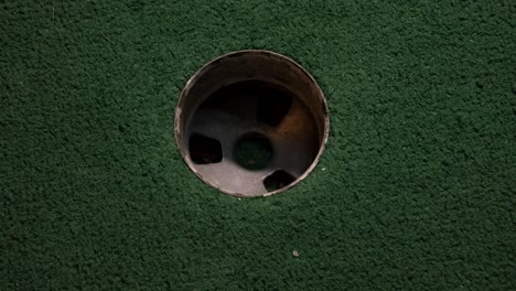 a close up birds eye view of a yellow mini golf ball falls into the golf hole and bounces in the pocket on a course