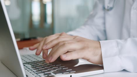 vista de primer plano de las manos del médico varón caucásico senior escribiendo en la computadora portátil en el escritorio