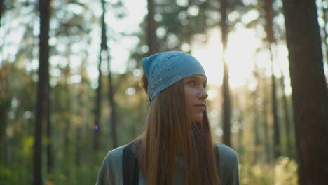 young woman wearing blue headscarf walking through peaceful forest, looking contemplative with soft sunlight creating a warm glow behind her, surrounded by trees and immersed in nature