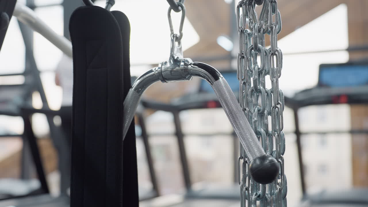 Silver curved grip handle and heavy chain attachment on cable machine in modern gym with blurred treadmills and large windows in background creating bright industrial fitness atmosphere