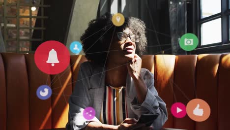 Animation-of-network-of-connections-with-icons-over-african-american-woman-using-technology