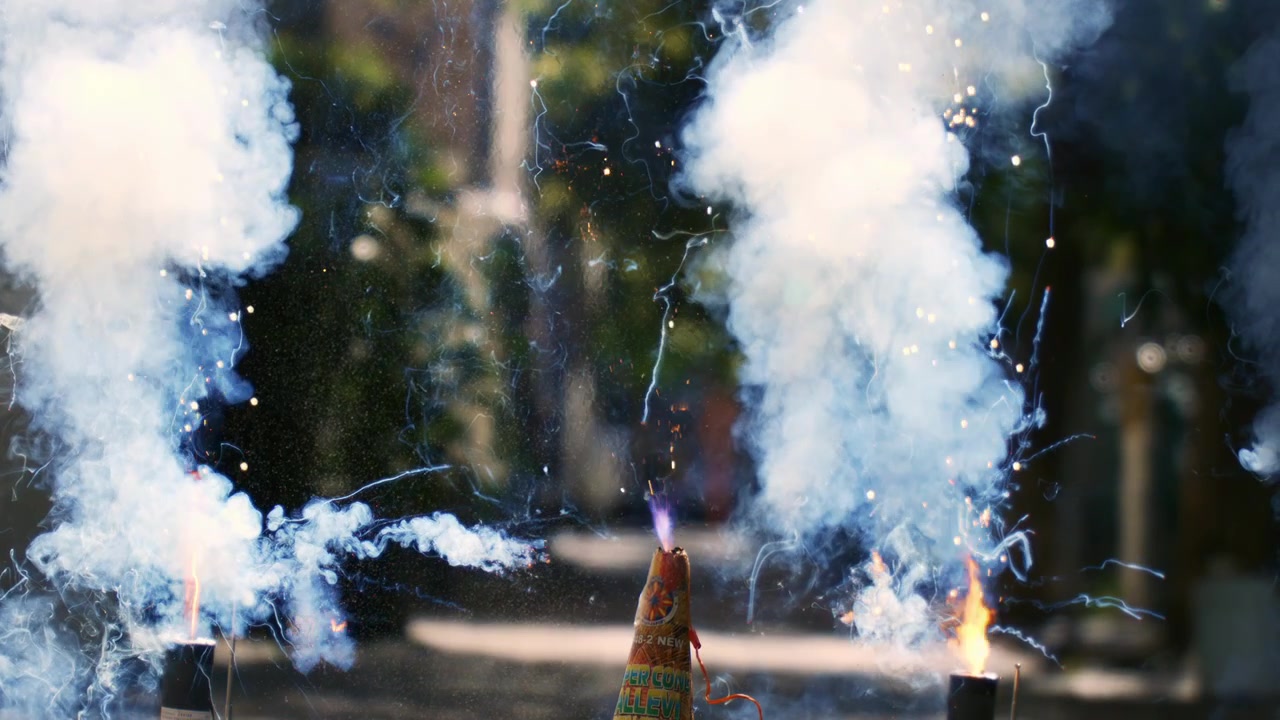 A fireworks cone ignites, emitting sparks and smoke on a street outdoors, showing the start of a pyrotechnic display.