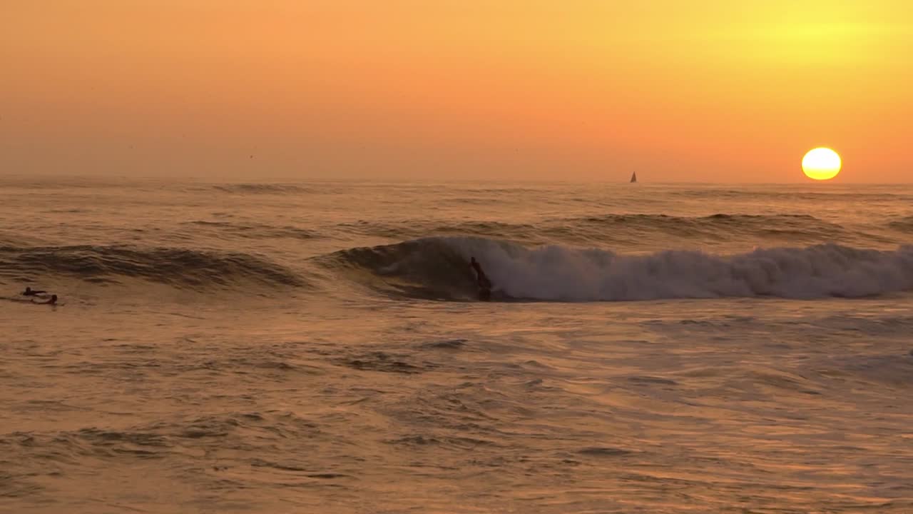 un tipo surfeando durante el atardecer con un velero en el fondo