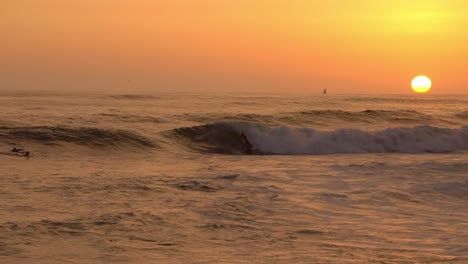 Chico-Surfeando-Durante-La-Puesta-De-Sol-Con-Un-Velero-En-El-Fondo