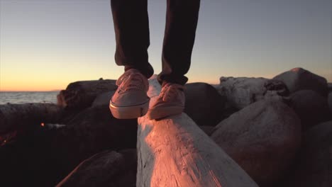 SLOW-MOTION:-Show-shot-of-man-walking-along-a-log-on-the-beach-by-the-ocean-at-sunset-4K