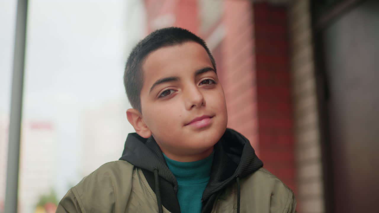 Kid in green jacket with soft warm smile looks at camera against blurred background of red brick building, exuding calm and friendly expression with natural outdoor light