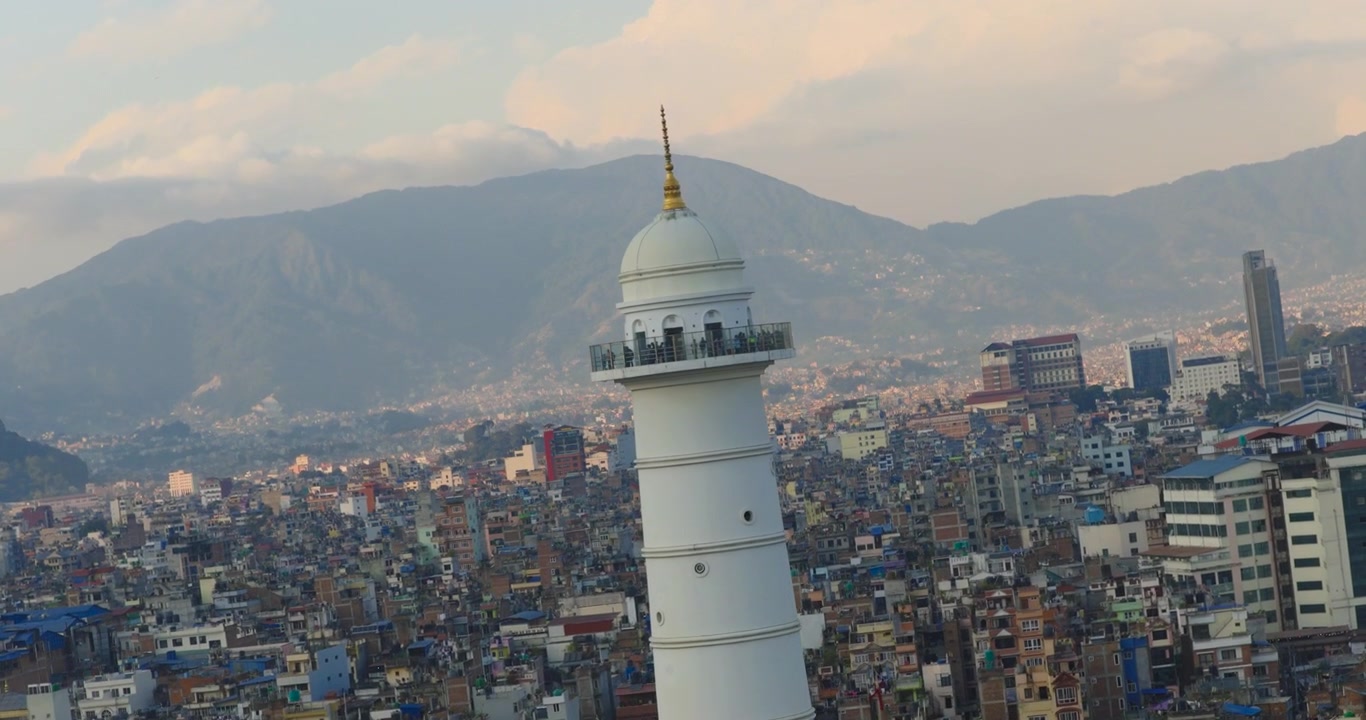 Dronella kuvattu otos Dharahara-tornista Kathmandussa, Nepalissa, paljastaa korkean valkoisen monumentin hallitsemattoman kaupunkimaiseman, vilkkaiden katujen ja ympäröivien laakson kukkuloiden keskellä, pehmeässä iltavalossa.
