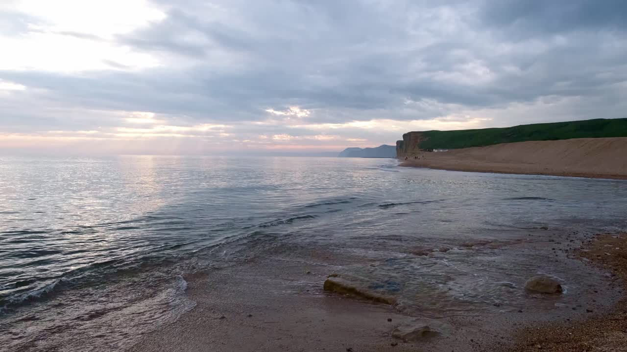 Jurassic Coast beach scene. Wide shot of the waves landing on the sand on a cloudy day. Cliff in the distance and sun peeking through over the horizon. Rocks and pebbles in the sand with white ripples