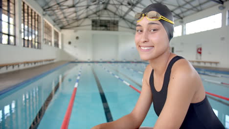 smiling female swimmer in swim cap and goggles sitting by indoor pool, copy space