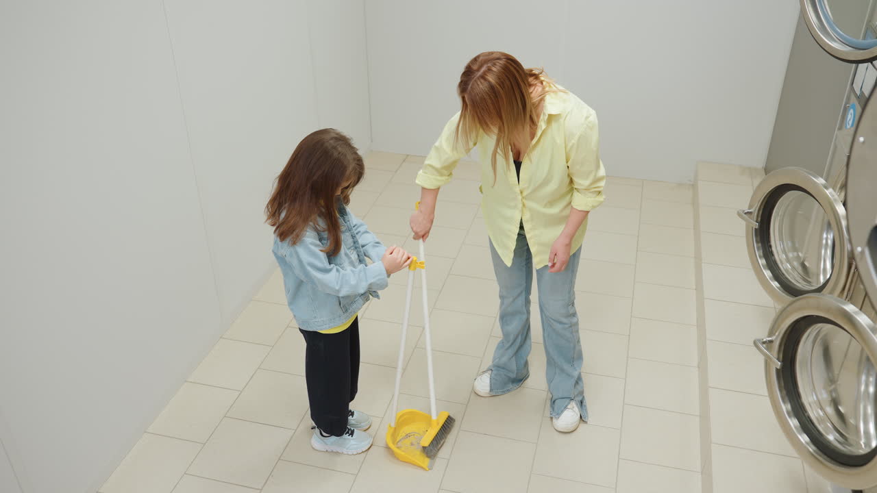 Overhead view shows shop owner sweeping tiled laundromat floor while toddler helper holds dustpan near stainless washers, teamwork routine, family hygiene focus, interior, denim jeans, yellow tools