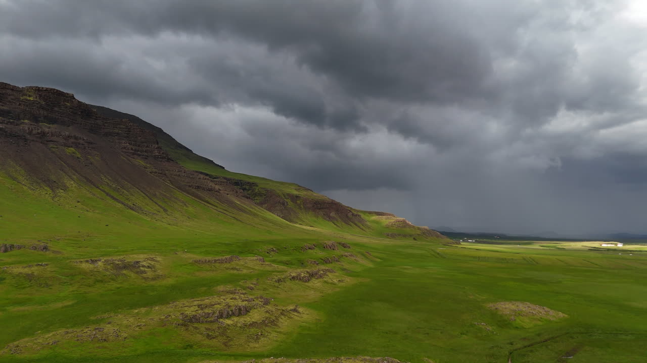 Wide aerial view of Snæfellsnes Peninsula showing shifting weather as rain clouds meet bright sunlight, revealing open green fields, distant mountains, and dramatic storm contrast across the landscape