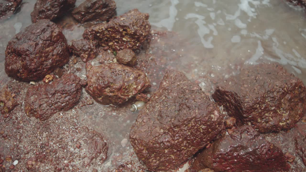 Detailed close-up shows reddish-brown laterite stones partially submerged at the shoreline, highlighting rough texture and mineral content on the coast of Goa, India