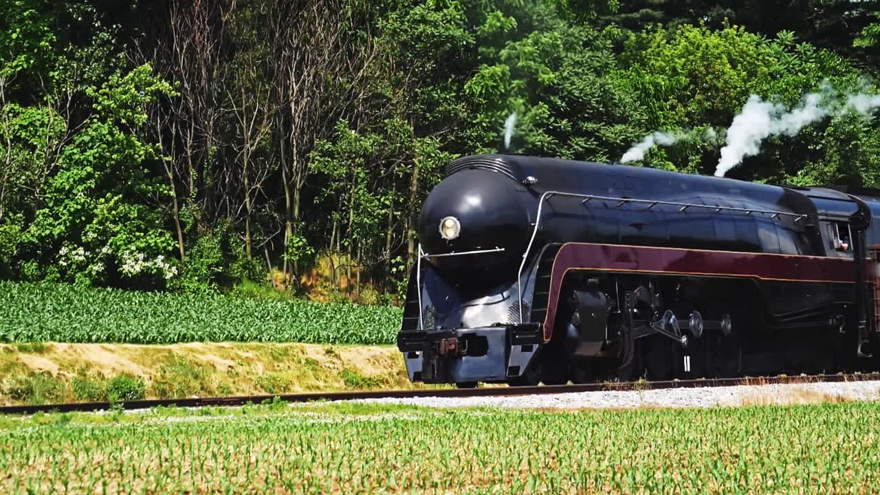 A classic steam locomotive chugs in slow motion along narrow tracks, surrounded by vibrant green fields. The warm sun casts a glow on the engine, creating a picturesque scene in the countryside.