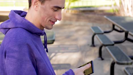 Un-Joven-Adulto-Navega-Por-Las-Redes-Sociales-Mientras-Habla-Por-Teléfono-En-Un-Parque,-Sonriendo