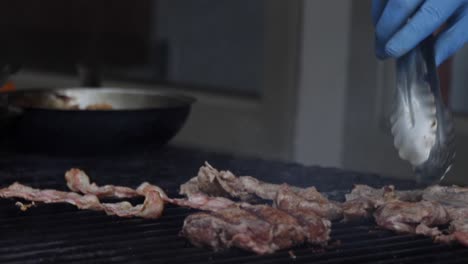 Slow-motion-shot-of-a-chef-turning-around-pieces-of-cooked-meat-and-bacon-on-the-grill