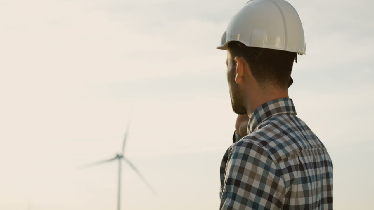 Side view of Caucasian man engineer wearing a helmet talking on the phone at wind station of renewable energy