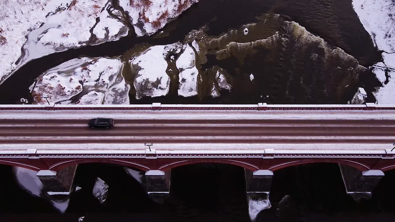 흐린 겨울날 라트비아 쿨디가의 벤타 강을 가로지르는 오래된 붉은 벽돌 다리의 공중 조감도, 배경의 벤타 급류, 뒤로 이동하는 광각 드론 샷