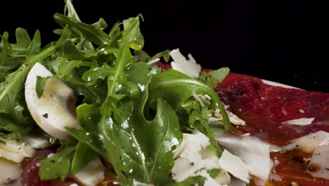 closeup moving over macro view beef carpaccio with rocket salad and slices of parmigiano falling from the sky 4k filmed on black background, italian food dish