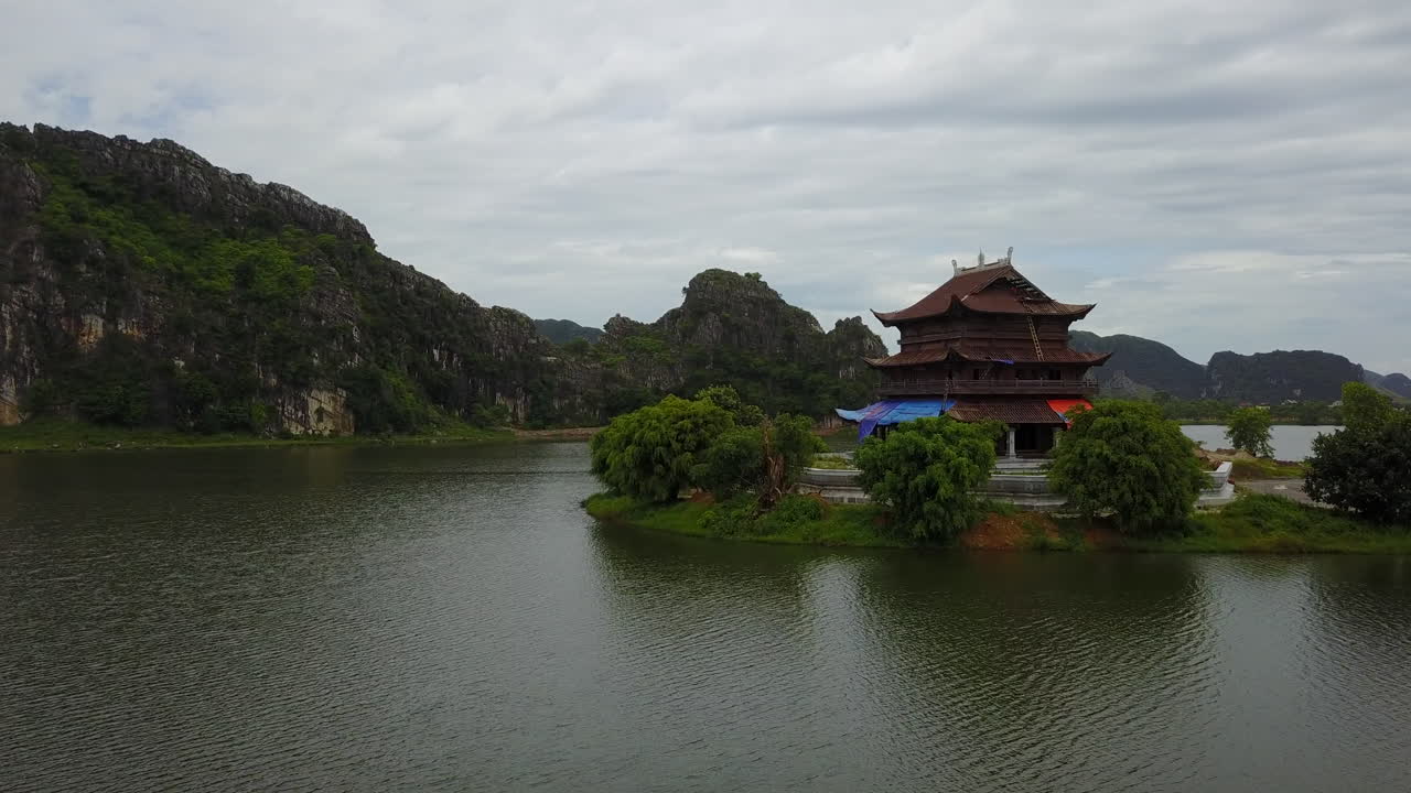 Experience an aerial view of the serene Đền thánh Cao Sơn temple in Tràng An, Ninh Bình, Vietnam, nestled amidst the region's stunning limestone karsts and waterways under a soft sky