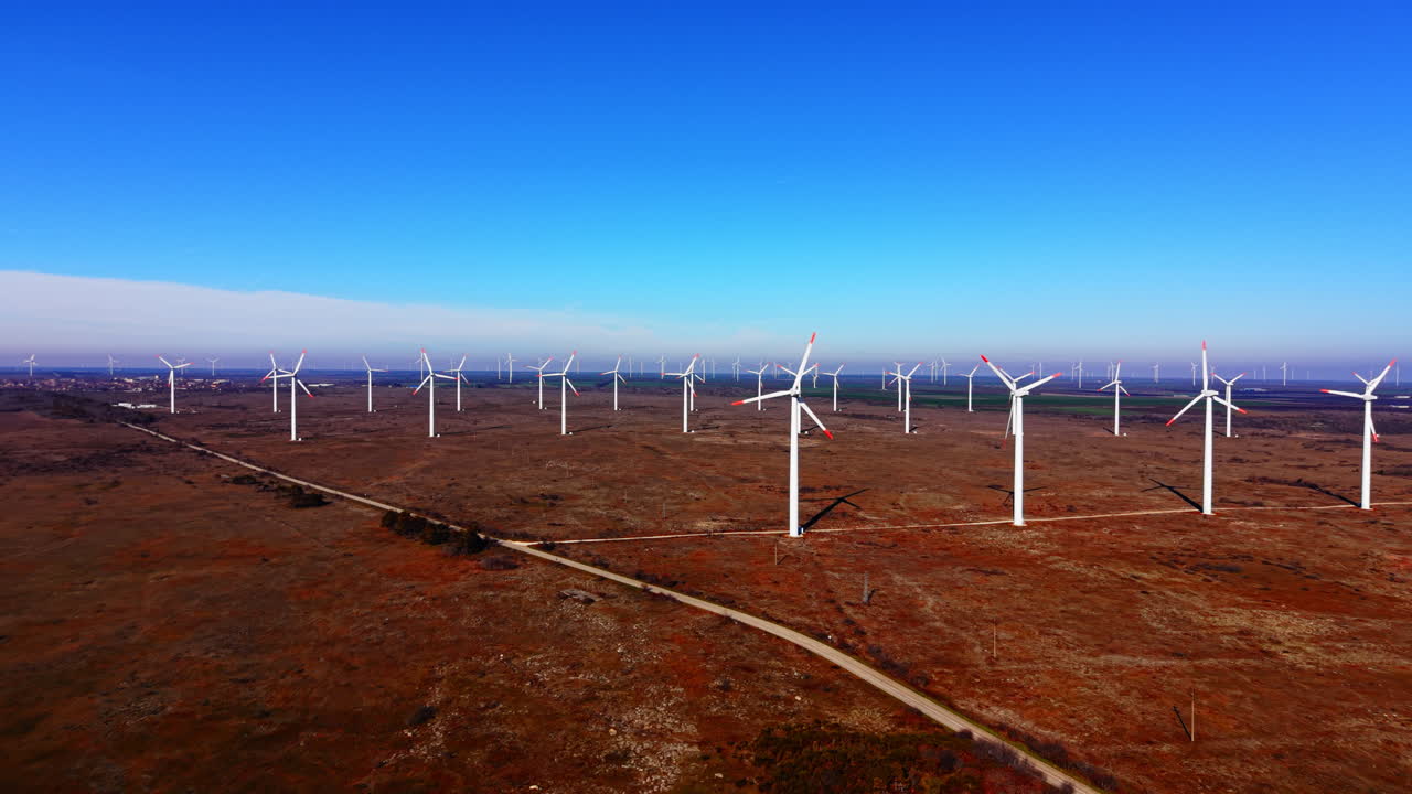 Vista aérea de alto ângulo de numerosos geradores eólicos espalhados por um vasto campo marrom com uma estrada de asfalto estreita cruzando a paisagem. Grande parque eólico em um campo rural com estrada.