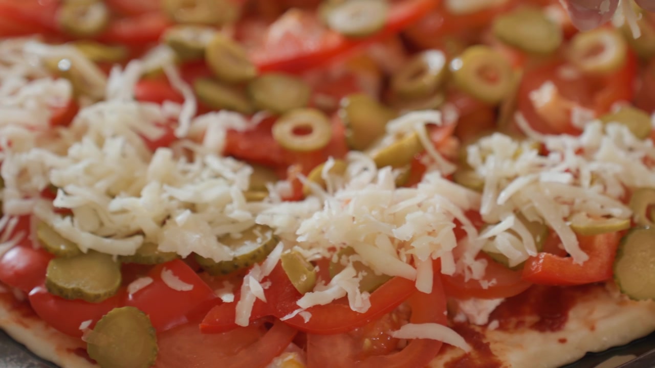 Vista ravvicinata dall'alto di una pizza rettangolare su un vassoio di metallo, coperta con fette di pomodoro e anelli di olive verdi mentre il formaggio grattugiato è sparso sulla superficie, mostrando la preparazione di un pasto saporito.