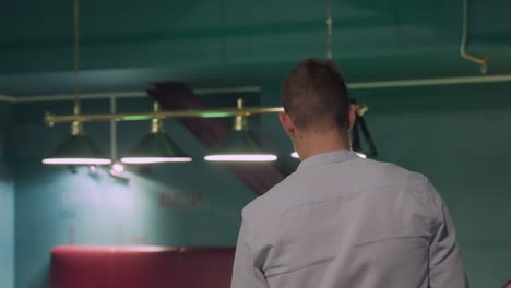 back shot of person in white shirt walking into billiard room with modern interior, overhead lamps illuminating pool tables. stylish setting with green walls, sleek furniture, and a relaxed ambiance