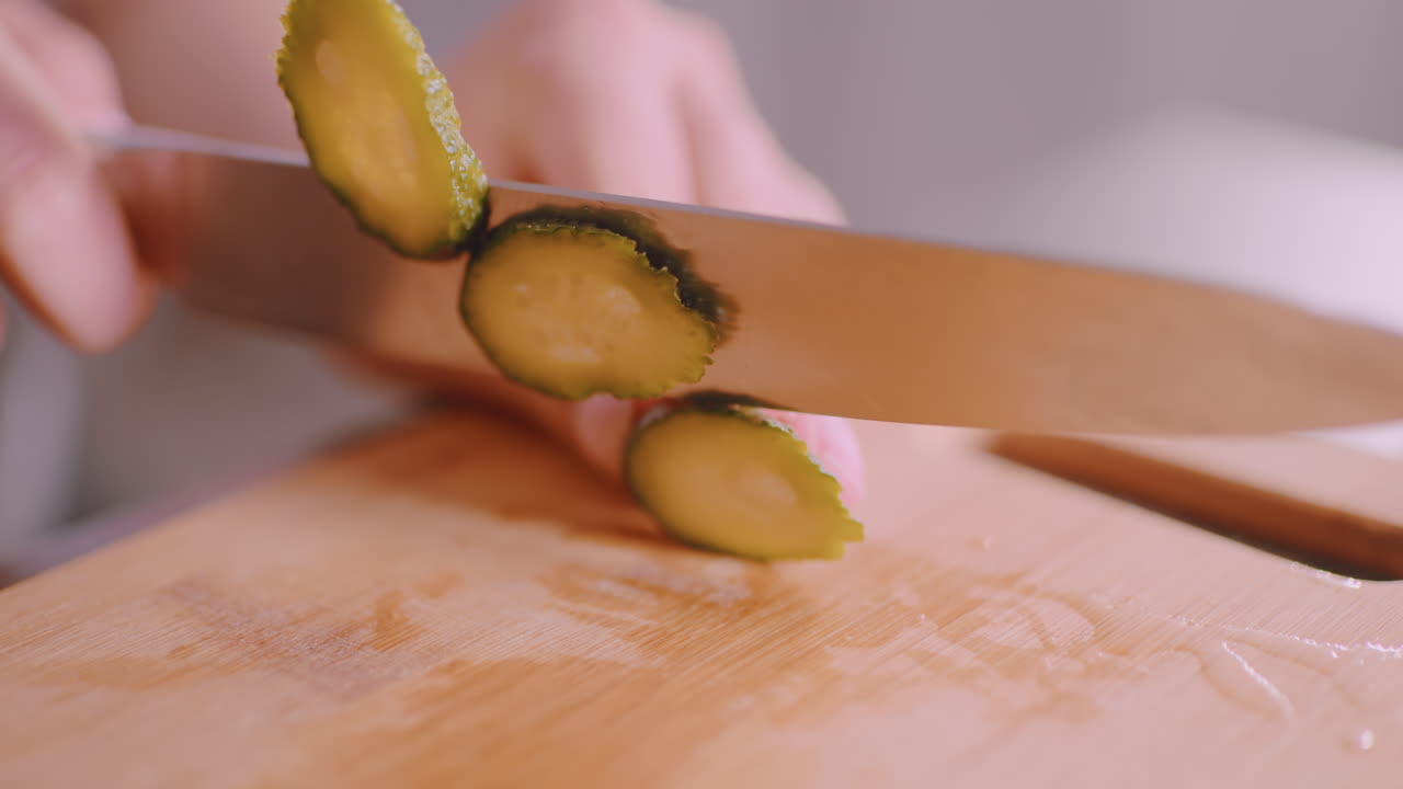 Close up of hand slicing fresh cucumber with sharp knife on wooden cutting board, showing detailed texture of vegetable and precision of cut during meal preparation in bright kitchen environment