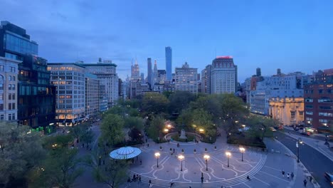 Ein-Zeitraffer-Des-Union-Square-Parks-In-Der-Abenddämmerung,-Der-Den-Übergang-Vom-Tag-Zur-Nacht-Festhält,-Mit-Den-Aufleuchtenden-Lichtern-Der-Stadt,-Menschen,-Die-Durch-Den-Park-Gehen,-Und-Der-Skyline-Von-New-York-Im-Hintergrund.