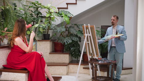 mujer posa para el famoso retratista en el estudio. dama glamurosa en vestido rojo con rosa y maestro de dibujo en la residencia. modelo gracioso con pintor