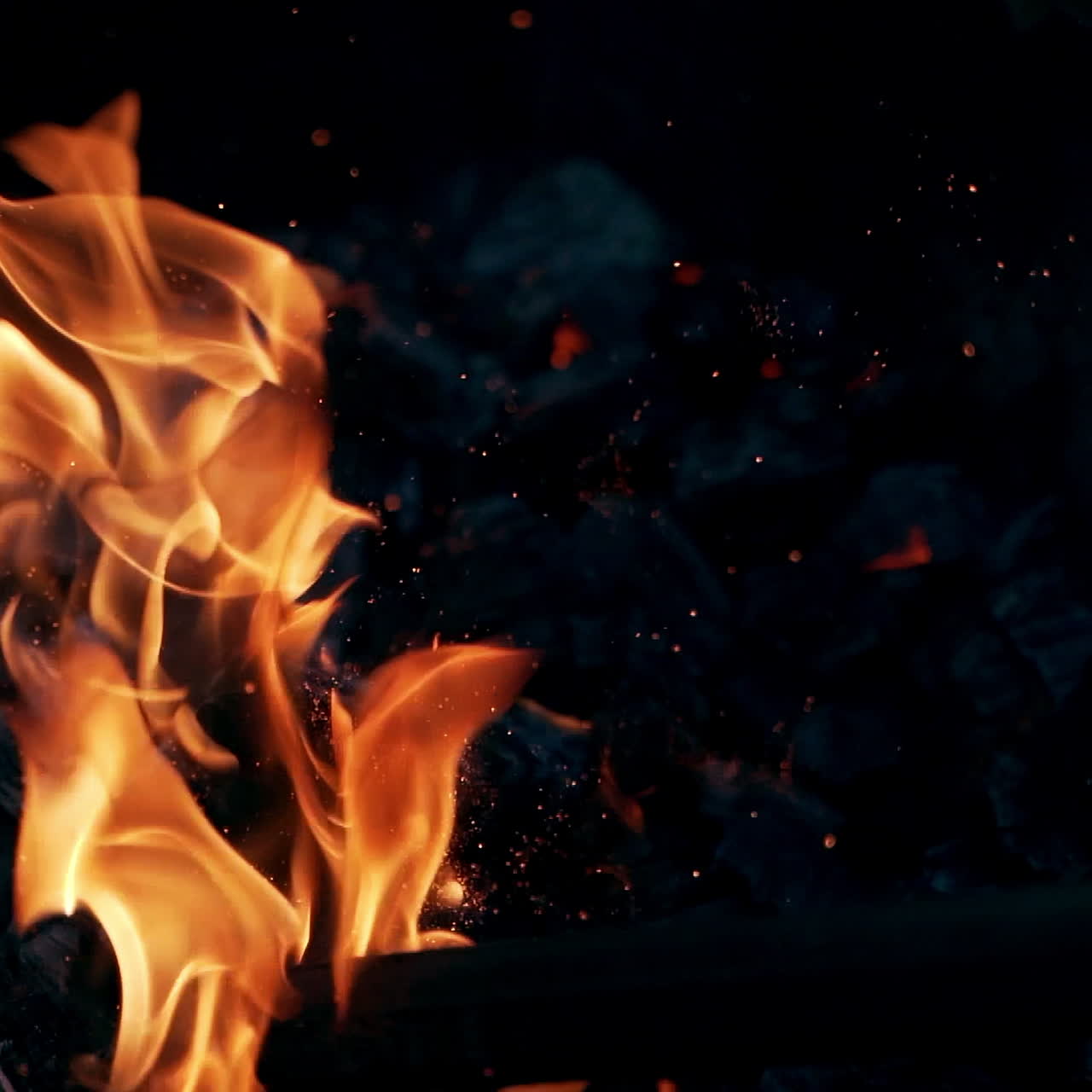 Wooden stick breaks smoldered logs in fire. Bright sparks moving on dark background from glowing fire. Slow motion.