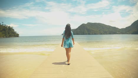 Una-Chica-Con-Un-Vestido-Corto-Sale-De-Una-Cabaña-Por-El-Embarcadero-Con-Olas-Rompiendo-En-La-Costa-Con-Montañas-Al-Fondo